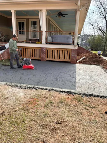 Worker compacting paver base with plate compactor beside a craftsman home with covered porch and brick columns