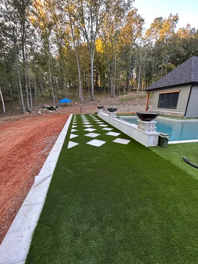 Artificial turf with alternating white stone stepping stones alongside a pool with decorative fire bowls